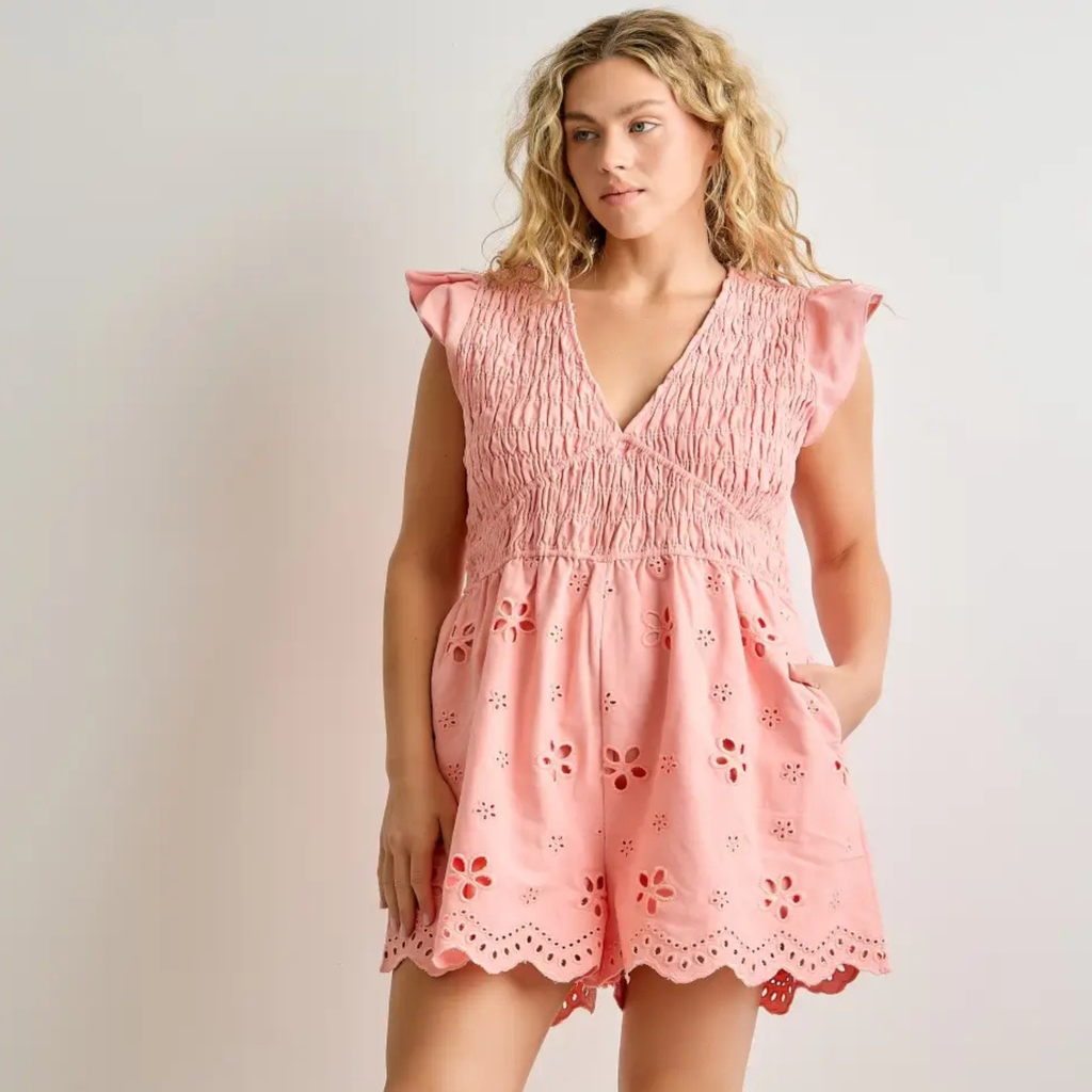 A woman modeling a light pink eyelet romper with a smocked bodice, ruffled cap sleeves, and a scalloped hem with floral cutout details, set against a plain light gray studio background.