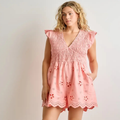 A woman modeling a light pink eyelet romper with a smocked bodice, ruffled cap sleeves, and a scalloped hem with floral cutout details, set against a plain light gray studio background.