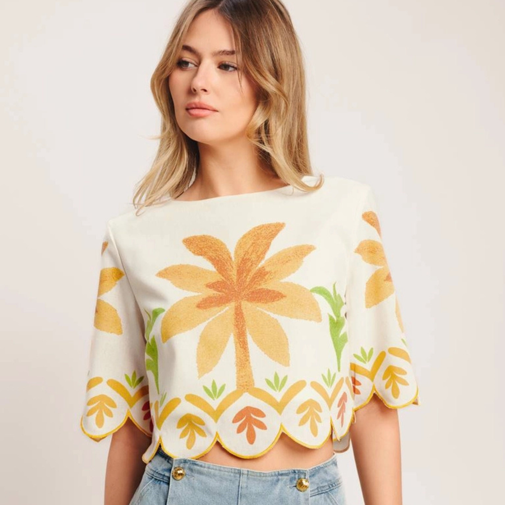 A woman wearing a cream-colored short-sleeve crop top with a large yellow palm tree and green leaf pattern, paired with light blue jeans, against a plain light background.