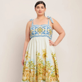 A woman modeling a sleeveless midi dress with a blue and yellow geometric patterned bodice and a cream skirt featuring a yellow palm tree and house print, set against a plain beige background.