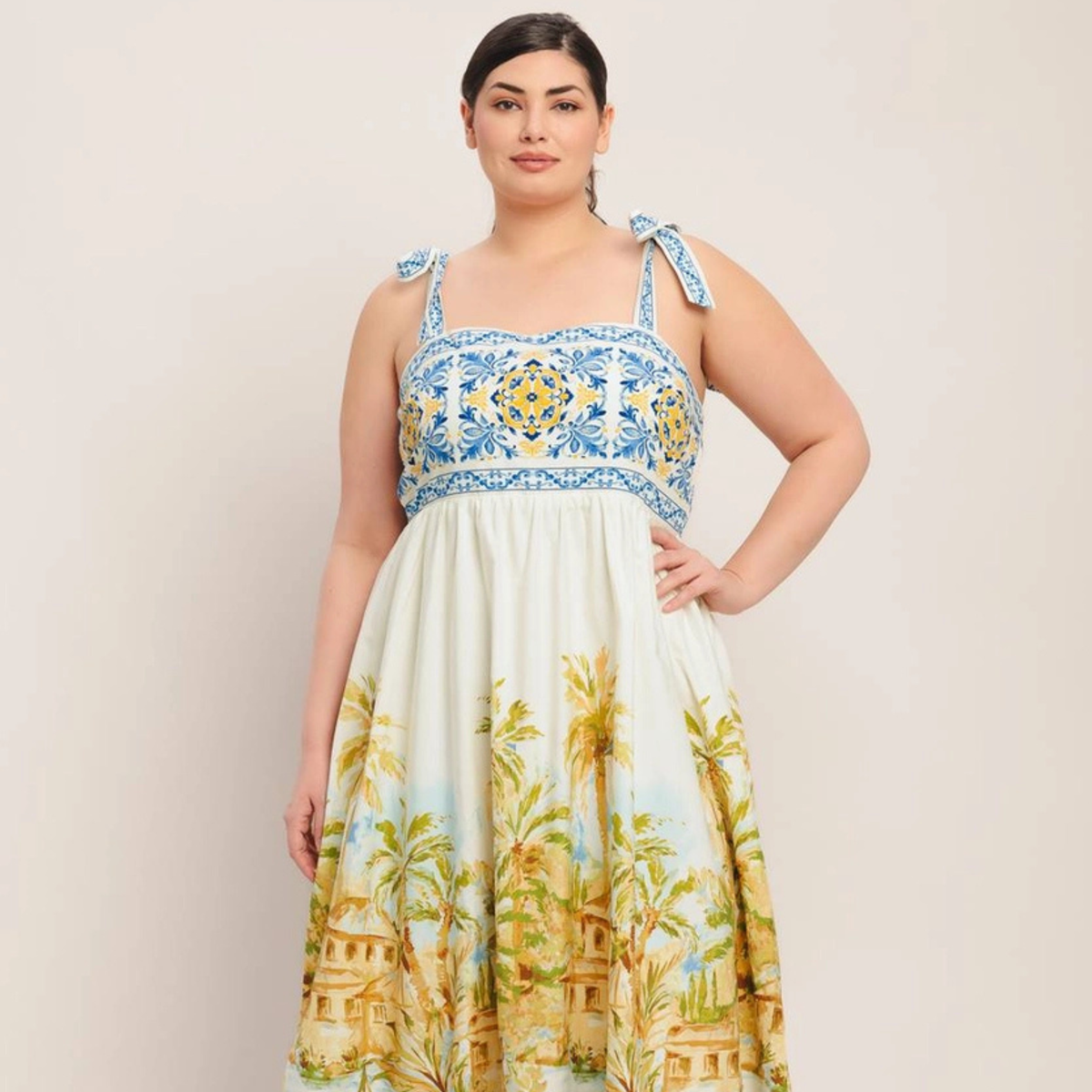 A woman modeling a sleeveless midi dress with a blue and yellow geometric patterned bodice and a cream skirt featuring a yellow palm tree and house print, set against a plain beige background.