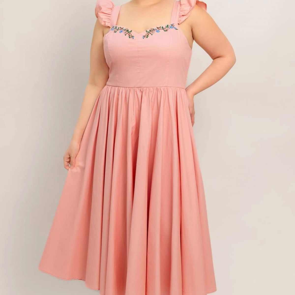 A woman wearing a light pink sleeveless midi dress with ruffled shoulder straps, a square neckline decorated with small blue and yellow floral embroidery, and a gathered flared skirt, photographed against a plain light gray background.