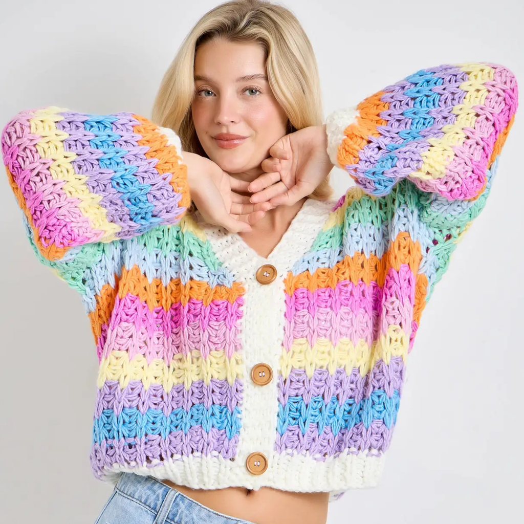 A woman wearing a chunky crocheted cardigan with horizontal rainbow stripes in pink, purple, blue, yellow, orange, and mint green, featuring a white V-neck trim and three wooden buttons.