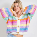 A woman wearing a chunky crocheted cardigan with horizontal rainbow stripes in pink, purple, blue, yellow, orange, and mint green, featuring a white V-neck trim and three wooden buttons.