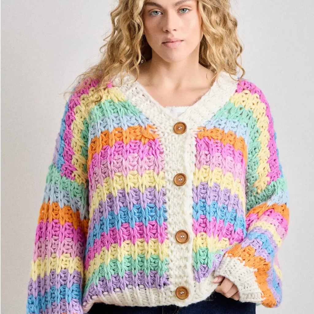 A woman wearing a chunky knit cardigan with horizontal rainbow stripes in pink, yellow, green, blue, purple, and orange, featuring a cream ribbed V-neck and button closure with brown wooden buttons.