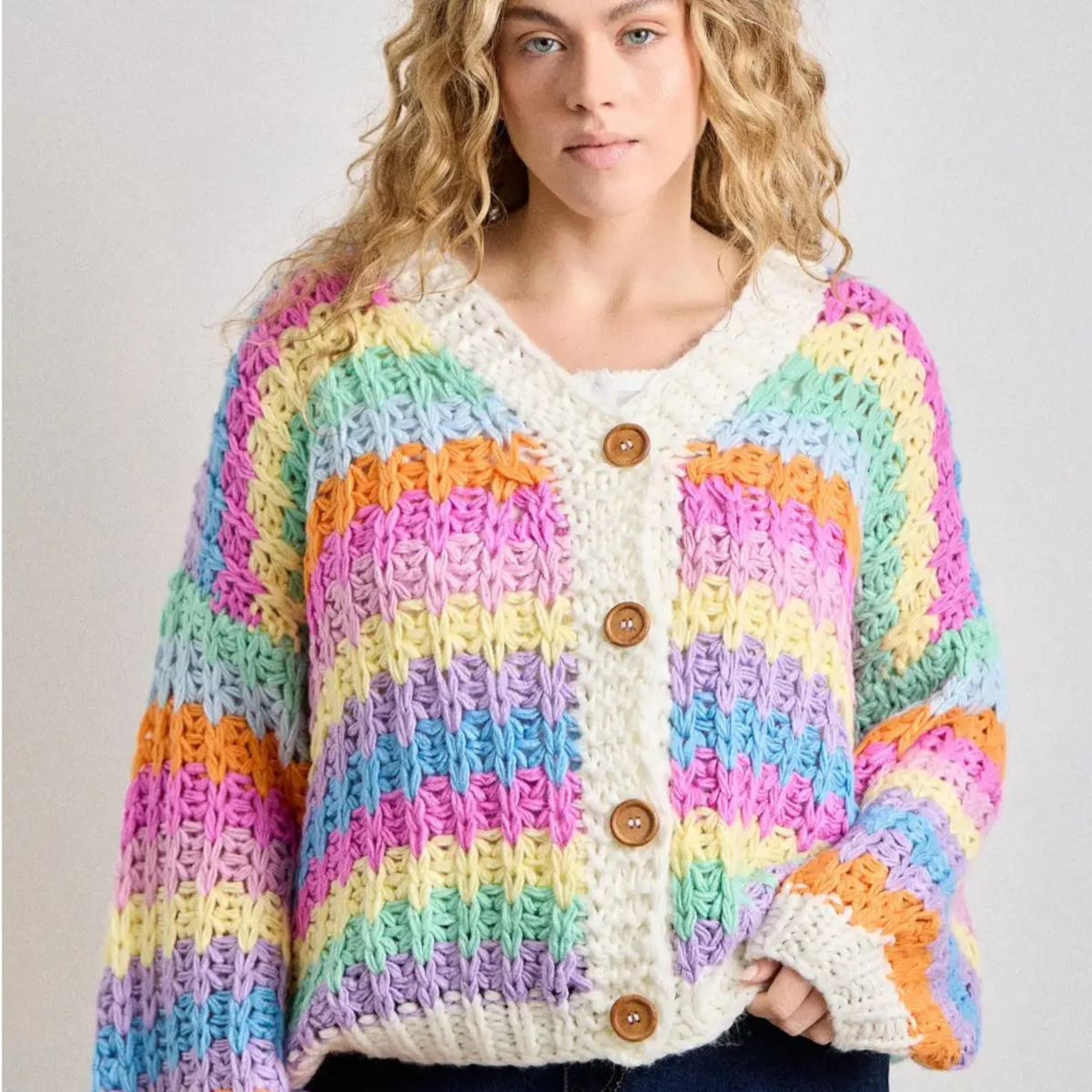A woman wearing a chunky knit cardigan with horizontal rainbow stripes in pink, yellow, green, blue, purple, and orange, featuring a cream ribbed V-neck and button closure with brown wooden buttons.