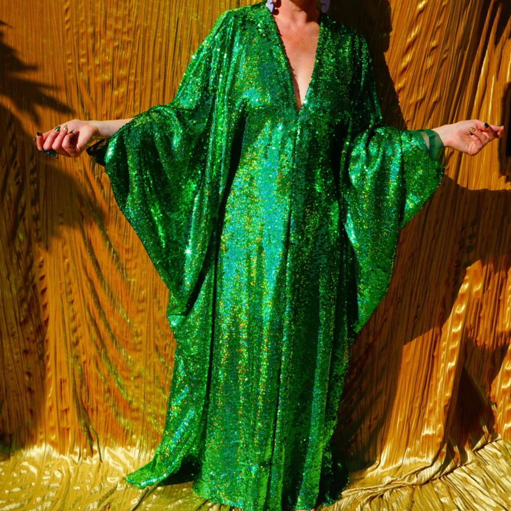 A person wearing a flowing, floor-length green sequined robe or caftan with wide draped sleeves, posed against a shiny gold textured backdrop.