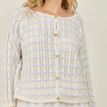 A close-up of a woman wearing a cream-colored knit cardigan with light blue wavy stripes and gold buttons, set against a plain light beige background.
