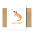 A white greeting card with a cartoon shrimp wearing a green party hat and the text 'HOPE YOUR BIRTHDAY IS SHRIMPLY AMAZING!' in orange, shown with a kraft paper envelope behind it on a white background.