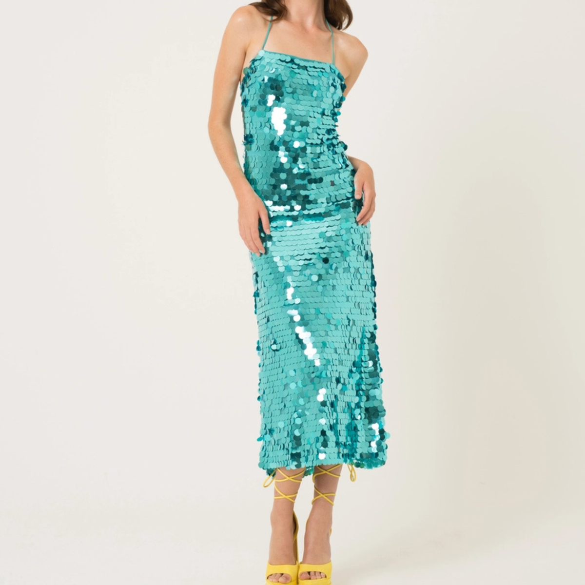 A woman modeling a turquoise sequined spaghetti-strap midi dress with yellow high-heeled sandals against a plain white studio background. Olivia Dean