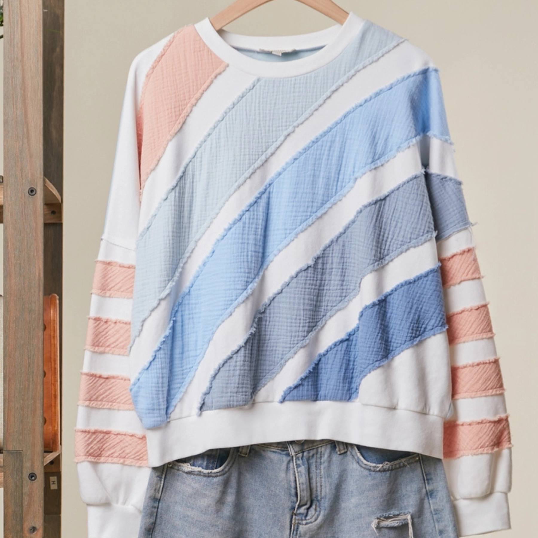 A white crewneck sweatshirt with diagonal textured stripes in light blue, medium blue, and soft pink, styled with light-wash denim jeans on a wooden hanger.