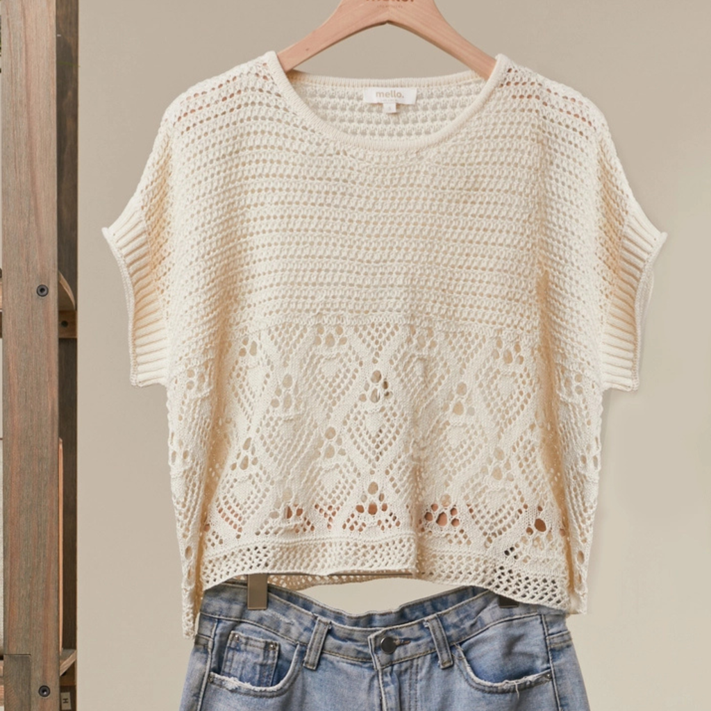 An off-white crochet knit short-sleeve crop top with a geometric openwork pattern, hanging on a wooden hanger above blue jeans.