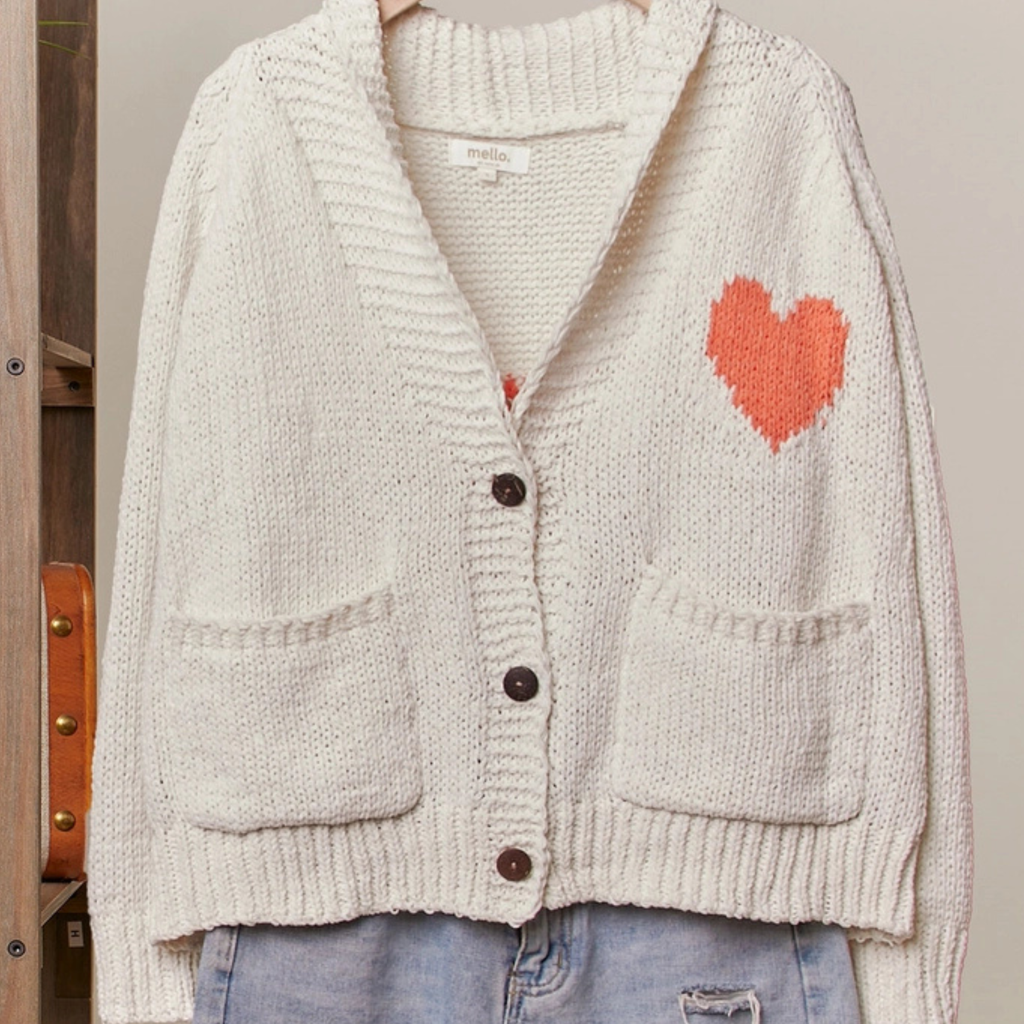A cream-colored knit cardigan sweater with a textured ribbed pattern, dark brown buttons, and a bright orange heart motif on the chest, styled with light blue jeans.