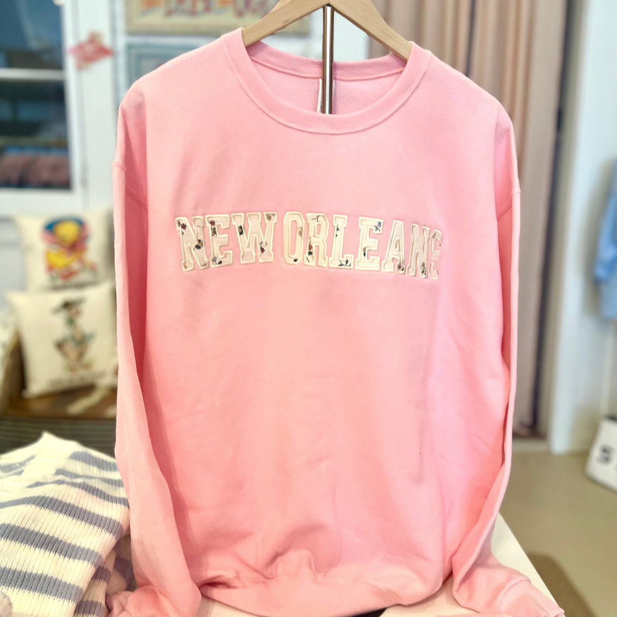 A pink crewneck sweatshirt hanging on a wooden hanger, featuring the word 'NEW ORLEANS' printed across the chest in a floral patterned font.
