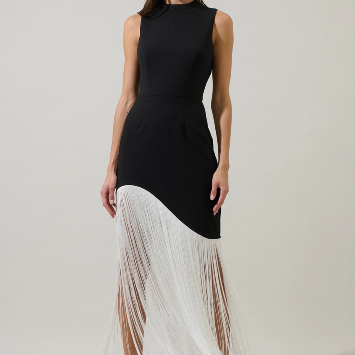 A sleeveless black dress with a high mock neck and a dramatic white fringe overlay on the lower half, shown on a model against a plain light beige studio background.
