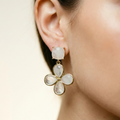 A close-up of a model's ear wearing a gold drop earring with a square white stone on top and a four-petal flower drop below, each petal featuring a milky white stone in a gold setting.
