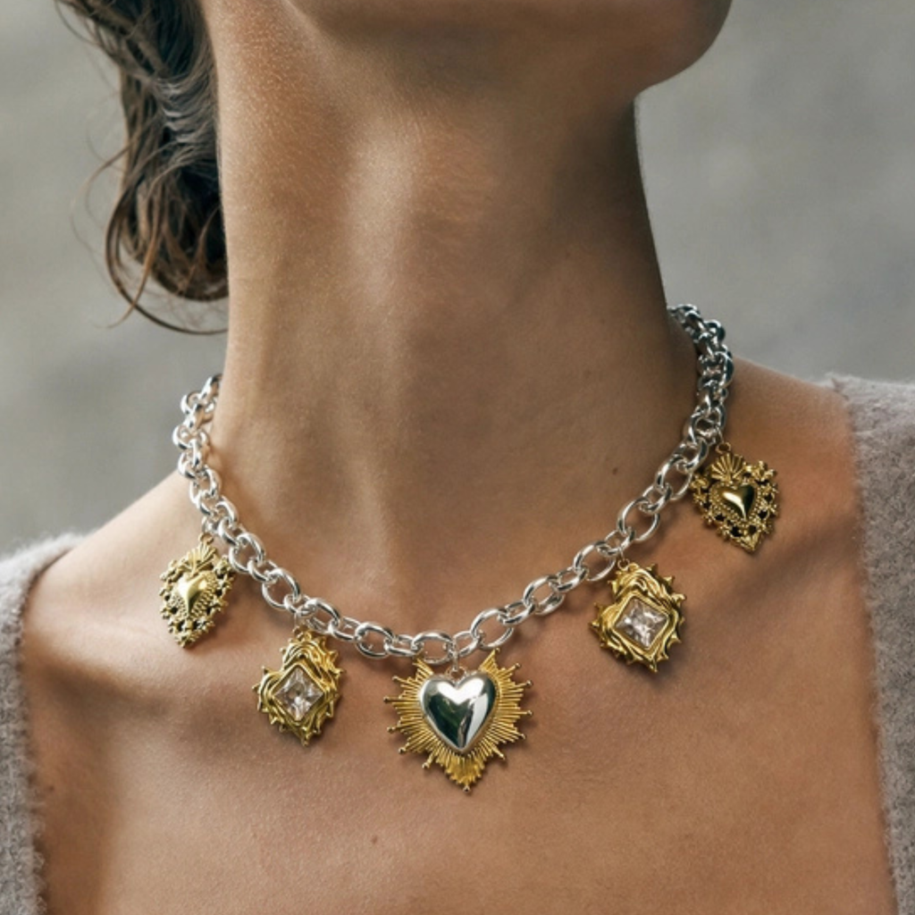 A close-up of a model's neck and collarbone wearing a silver chain necklace adorned with five ornate gold heart-shaped pendants, including one larger central heart pendant.