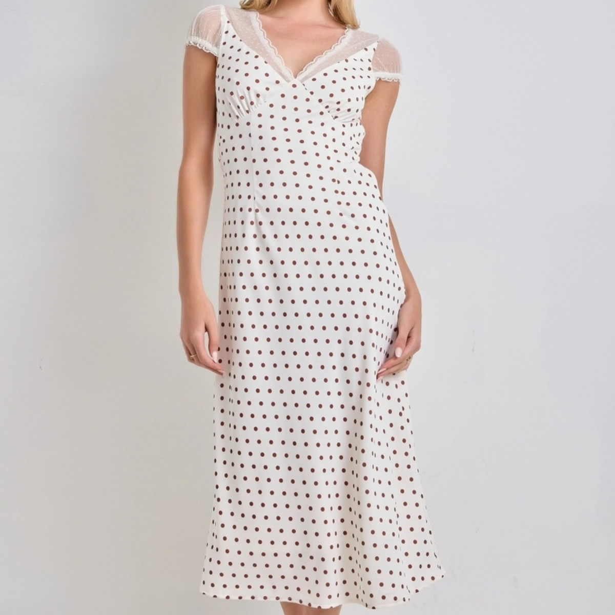 A woman wearing a cream-colored midi dress with a brown polka dot pattern, sheer cap sleeves, and a lace V-neckline, photographed against a plain light gray background.