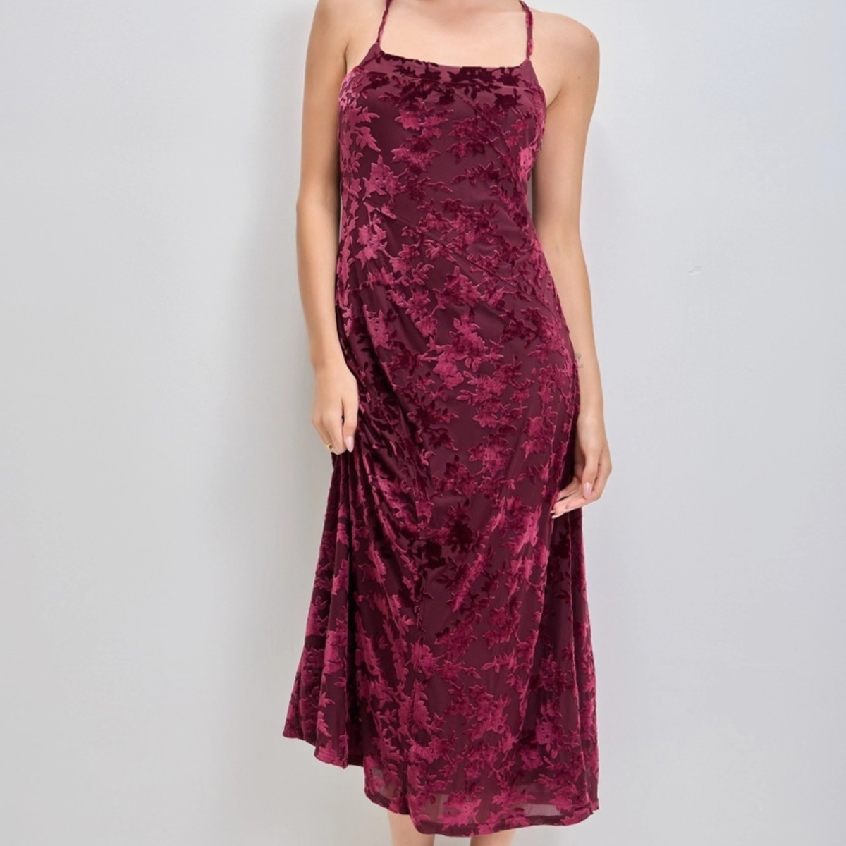 A woman wearing a sleeveless midi dress made of crushed velvet in a deep burgundy color, shown against a plain light gray background.