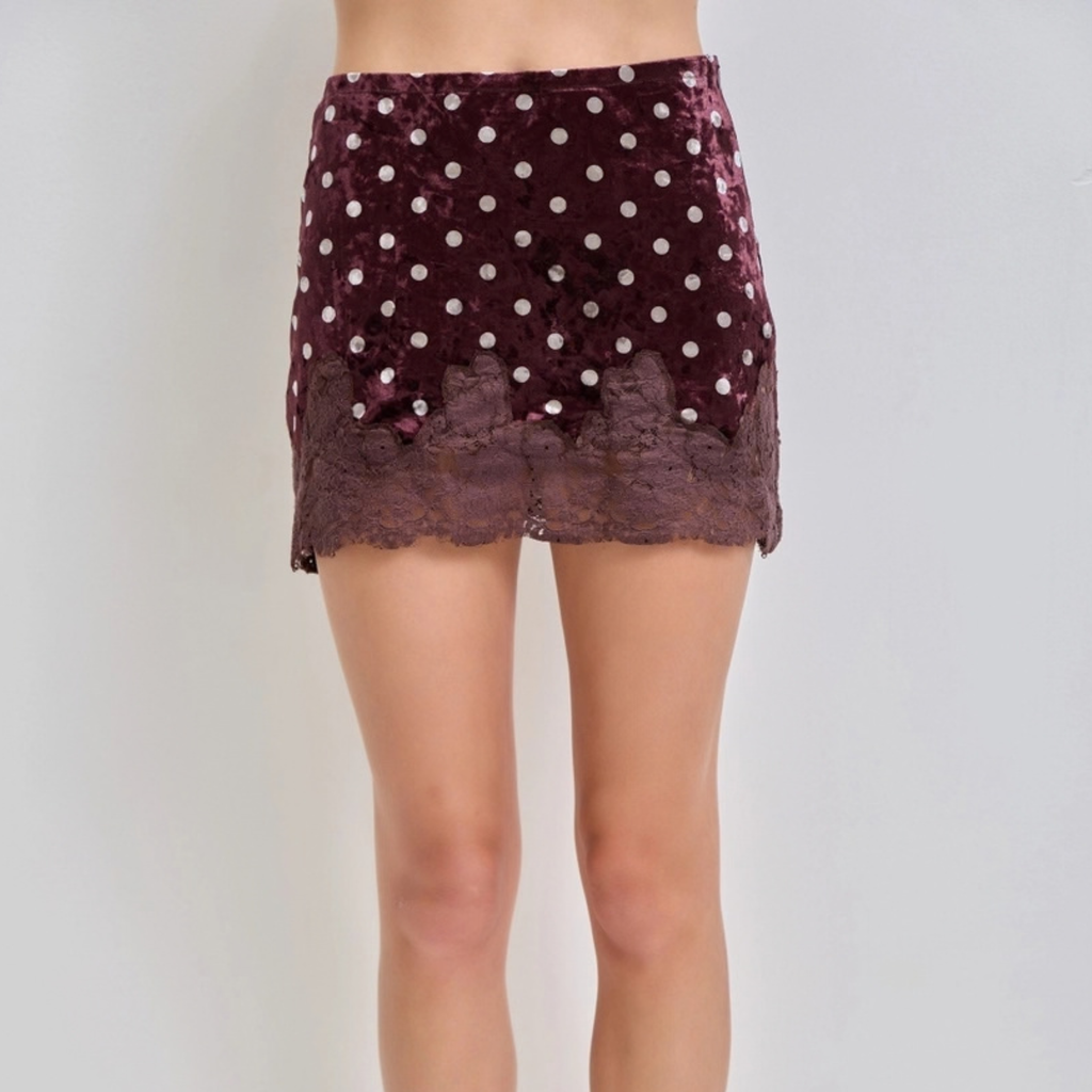 A model's lower body wearing a short velvet mini skirt in a deep plum purple with white polka dots and a matching lace hem, set against a plain light gray background.