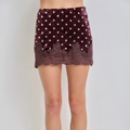 A model's lower body wearing a short velvet mini skirt in a deep plum purple with white polka dots and a matching lace hem, set against a plain light gray background.