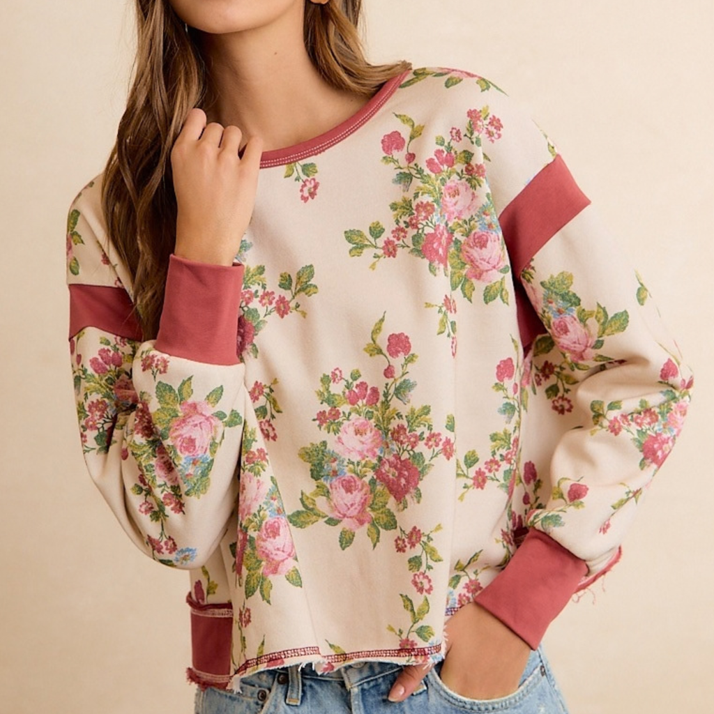 A woman wearing a cream-colored long-sleeve sweatshirt with a pink and green floral print and dusty rose ribbed trim, paired with blue jeans against a warm beige background.