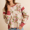 A woman wearing a cream-colored long-sleeve sweatshirt with a pink and green floral print and dusty rose ribbed trim, paired with blue jeans against a warm beige background.