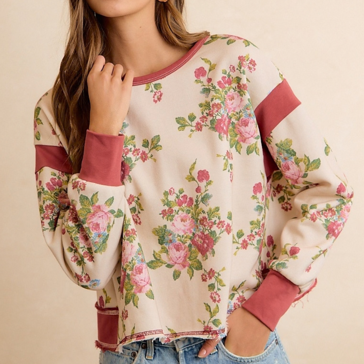 A woman wearing a cream-colored long-sleeve sweatshirt with a pink and green floral print and dusty rose ribbed trim, paired with blue jeans against a warm beige background.