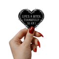 A hand with red manicured nails holds a heart-shaped black sticker with white text reading 'LIFE'S A BITCH, THANKFULLY SO AM I' and a white barbed wire border, against a plain white background.