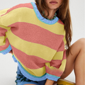 A close-up of a model wearing a colorful knit sweater with wide horizontal stripes in pastel yellow, dusty pink, and light blue, paired with blue patterned shorts.