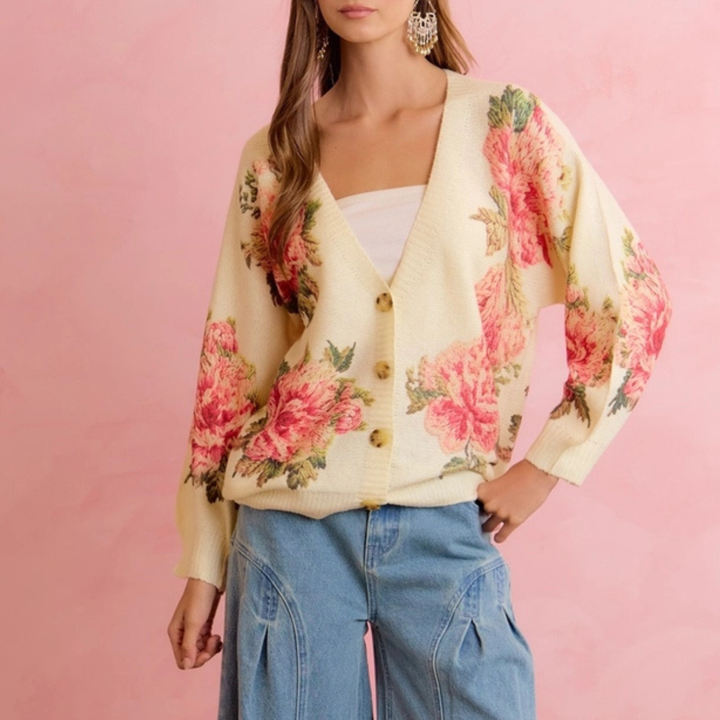 A woman wearing a cream-colored cardigan with a vibrant pink and red floral print, styled with light blue jeans against a solid pink background.