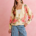 A woman wearing a cream-colored cardigan with a vibrant pink and red floral print, styled with light blue jeans against a solid pink background.