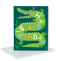 A green crocodile birthday card with colorful letters spelling 'HAPPY BIRTHDAY' and a small red party hat, shown standing upright with a white envelope behind it on a white background.