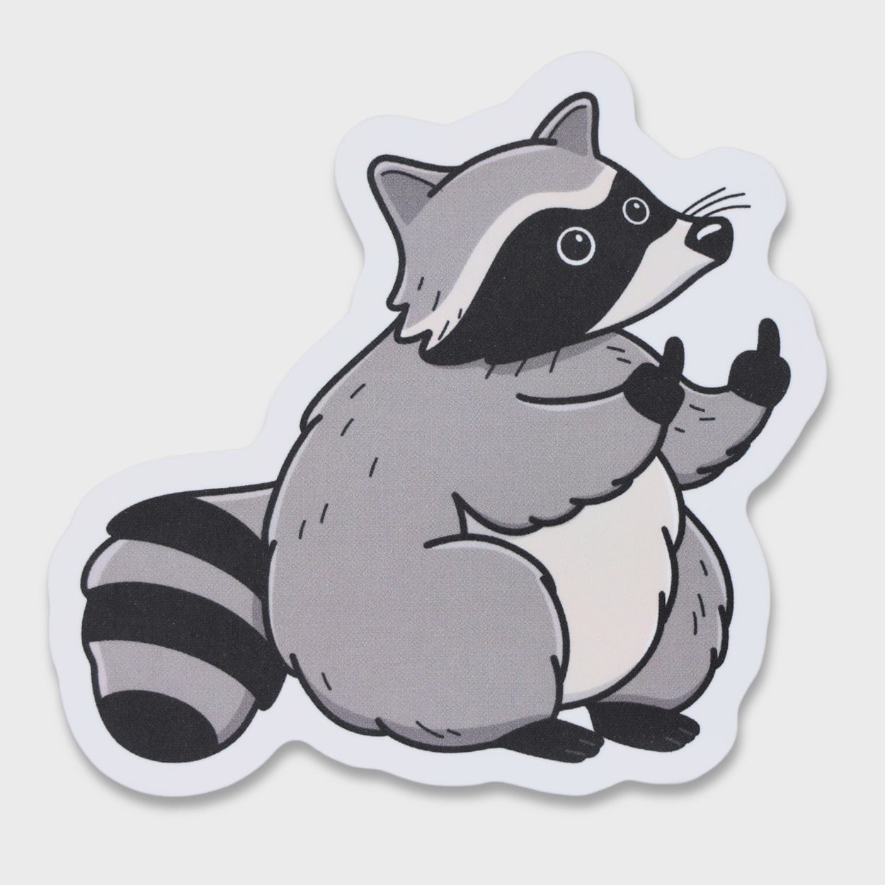 A die-cut sticker featuring a cartoon raccoon in gray, black, and white, shown on a light gray background.
