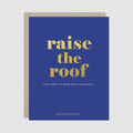A blue birthday card with gold and white text reading 'raise the roof' and 'HAPPY BIRTHDAY!', shown with a beige envelope behind it on a light gray background.