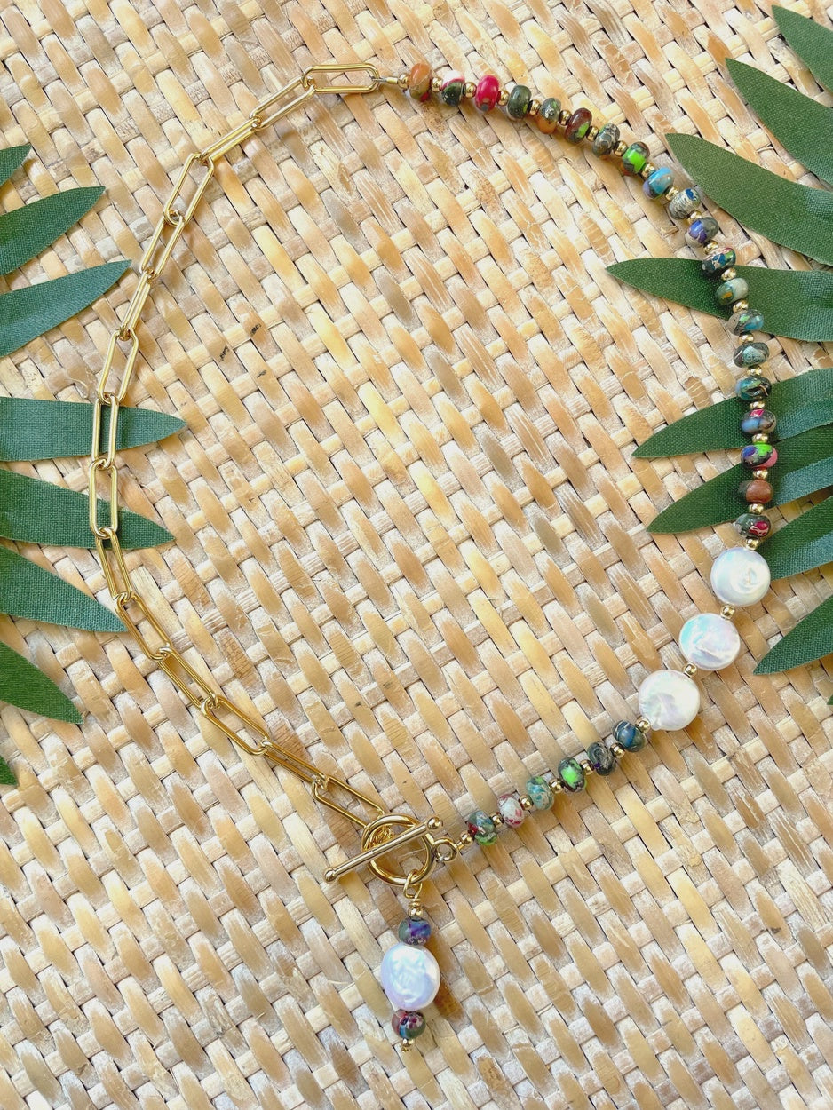 A gold chain necklace with a toggle clasp, decorated with small multicolored beads and white baroque-style shell beads, laid flat on a woven straw mat with green leaves.
