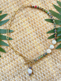A gold chain necklace with a toggle clasp, decorated with small multicolored beads and white baroque-style shell beads, laid flat on a woven straw mat with green leaves.