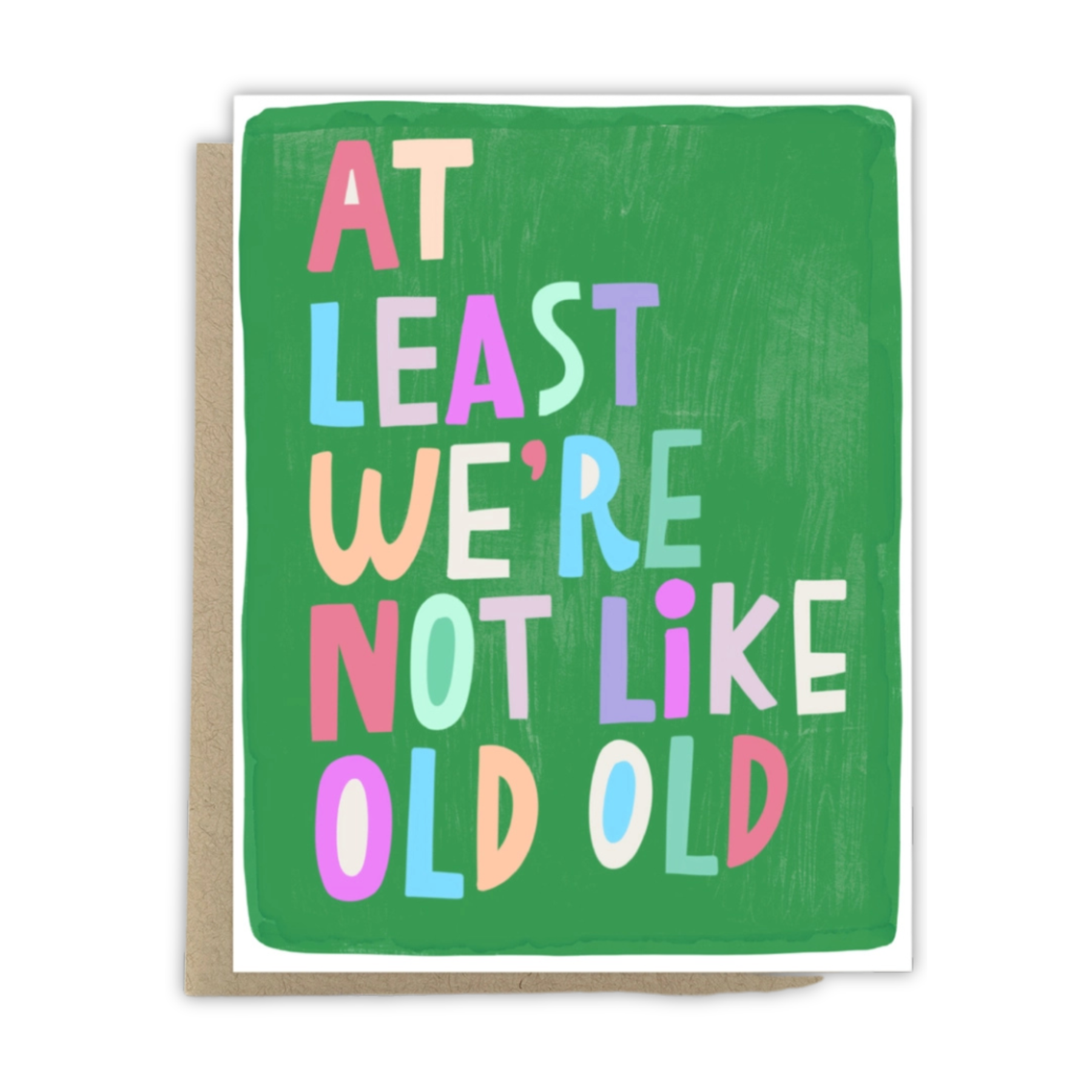 A square greeting card with a green textured background and colorful hand-painted text reading 'AT LEAST WE'RE NOT LIKE OLD OLD'. The card is shown with a kraft paper envelope behind it on a white background.