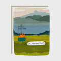 A square greeting card with a humorous landscape illustration. The scene shows a campfire burning in a grassy field with mountains and water in the background.
